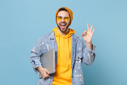 Funny young hipster guy in fashion jeans denim clothes posing isolated on pastel blue background in studio. People lifestyle concept. Mock up copy space. Hold laptop pc computer, showing OK gesture.