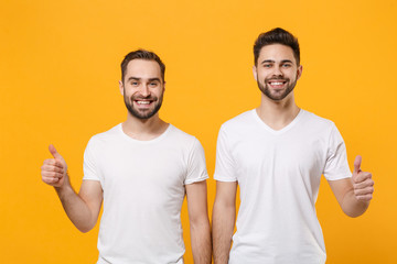Smiling young men guys friends in white blank empty t-shirts posing isolated on yellow orange background studio portrait. People emotions lifestyle concept. Mock up copy space. Showing thumbs up.