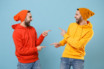 Two laughing hipster men guys in fashion red yellow clothes posing isolated on pastel blue background studio portrait. People lifestyle concept. Mock up copy space. Point index fingers at each other.