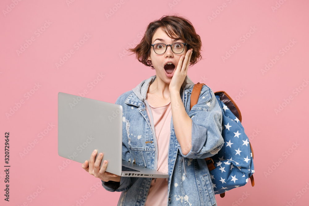 Poster Shocked young woman student in denim clothes glasses backpack isolated on pastel pink background. Education in high school university college concept. Working on laptop pc computer put hand on cheek.