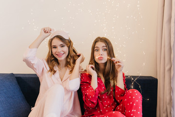 Good-looking young ladies in pajamas posing on couch with lightbulbs on background. Indoor shot of lovable sisters chilling on blue sofa.