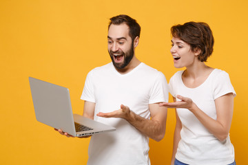 Excited young couple friends bearded guy girl in white blank empty t-shirts isolated on yellow orange background. People lifestyle concept. Mock up copy space. Pointing hands on laptop pc computer.