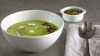 Broccoli soup served with some pumpkin and sunflower seeds, fresh cream, olive oil, black pepper and parmesan cheese in a white bowl. There is a spoon on a napkin and some seeds in little bowl near it