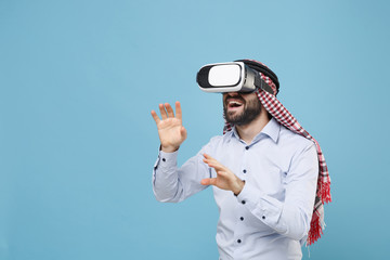 Funny arabian muslim man in keffiyeh kafiya ring igal agal casual clothes isolated on pastel blue background. People religious lifestyle concept. Watch in headset touch something like push on button.