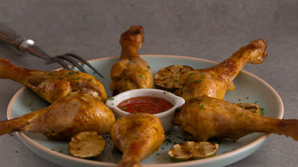 Six cooked chicken legs on a big blue round plate. Served with grilled lime and some sweet chilli sauce in a small white bowl. View from the side. There is big chicken fork in a plate. Grey background