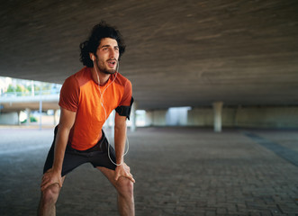 Portrait of a tired caucasian athlete man listening to music taking breathe after a long run under...