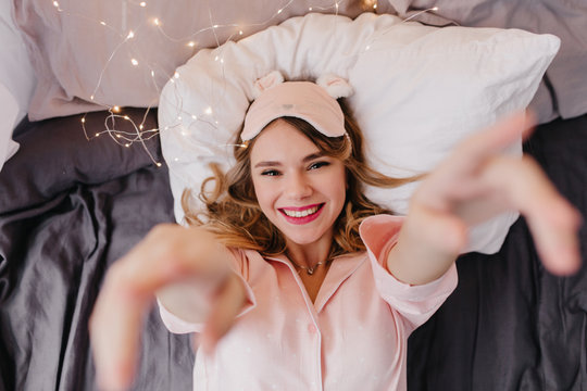 Pleased blonde girl lying on dark sheet. Indoor overhead photo of smiling cheerful lady in pink eyemask posing in bed.