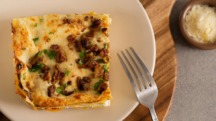 Butternut  vegetarian lasagne in a plate on a wooden board. With chopped nuts and herbs on it. View from the top. There is a fork in a plate and some parmesan cheese in little bowl. 