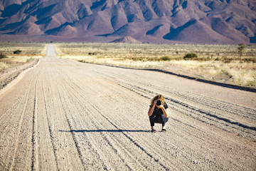 Young woman with a camera squatting in the middle of a gravel road that is leading into a...
