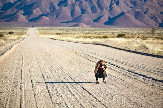 Young woman with a camera squatting in the middle of a gravel road that is leading into a mountainous landscape near Spreetshoogte Pass, Namibia, Africa