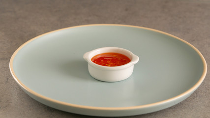 Big round blue plate with little white bowl with red sweet hot chilli sauce in it. Grey background