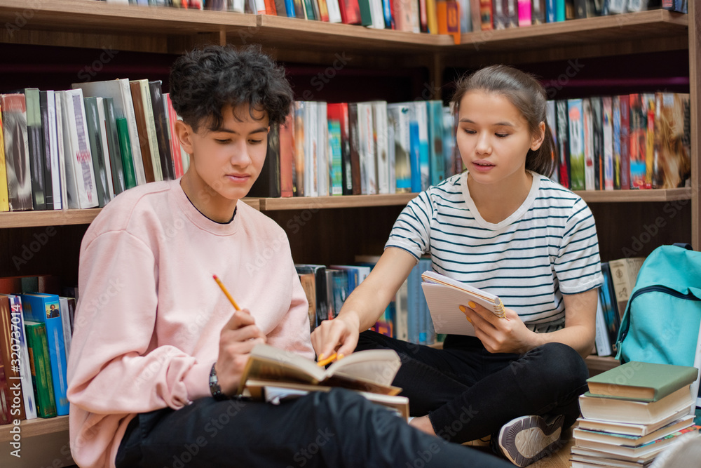 Poster Clever confident girl pointing at page of book held by her classmate during preparation for seminar in college library