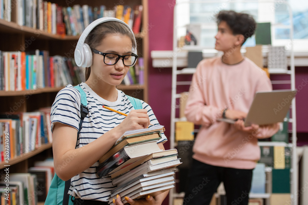 Poster Pretty girl in headphones and eyeglasses holding stack of books while standing by bookshelf and making list of literature