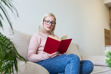 Young beautiful woman lying on sofa and reading book at home. Excellent book. Beautiful young woman reading a book at the sunny morning.