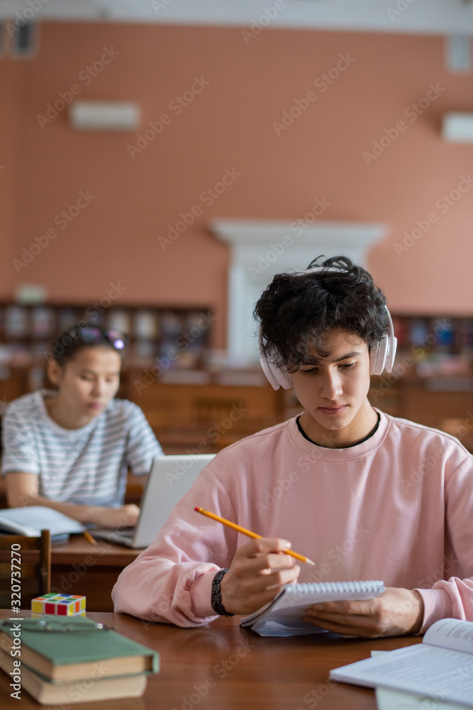 Poster Serious college student in headphones listening to music while reading notes in notepad by desk with girl on background