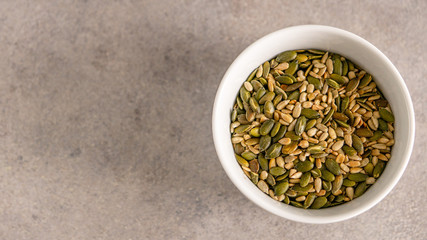 Pumpkin and sunflower seeds in white bowl based  on the right side. Grey background. 