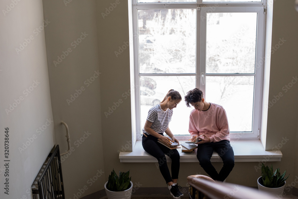Poster two youthful students in casualwear sitting on windowsill in college corridor while preparing home t