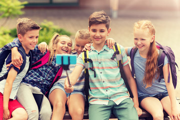 primary education, technology, friendship, childhood and people concept - group of elementary school students with backpacks sitting on bench and taking picture by smartphone on selfie stick outdoors