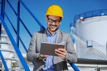 Handsome smiling unshaven businessman in suit and with helmet on head using tablet. In background are oil tanks.
