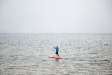 Athletic man standing with a paddle on the red surfboard at early misty morning. Stand up paddle boarding.