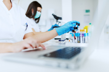 Female lab assistant using laptop for data entry and in other hand holding test tube with blood...