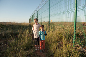 two poor kids brother and sister refugees standing in green grass near state border with long fence...