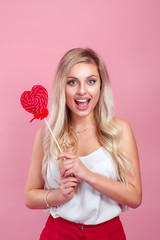 Blonde happy girl holding a red heart-shaped lollipop. Valentine's Day