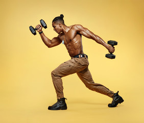 Sporty man training muscles of hands and legs using a dumbbells. Photo of man with good physique isolated on yellow background. Strength and motivation