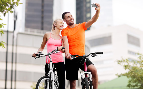 fitness, sport and people concept - happy couple with bicycles taking selfie by smartphone over city street on background