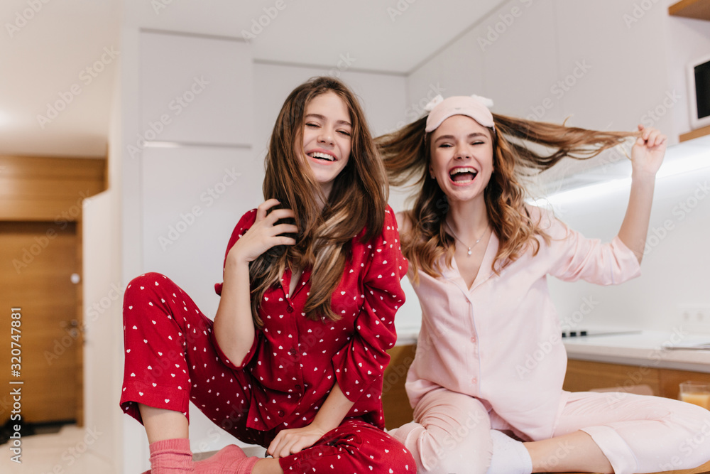 Wall mural Graceful white girl with curly hairstyle enjoying good morning with sister. Indoor shot of inspired caucasian female models in night-suits playing with their hair.