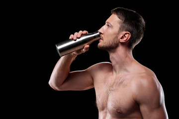 Fototapeta na wymiar Young shirtless athlete standing in front of camera and drinking water from metal bottle after workout over black background