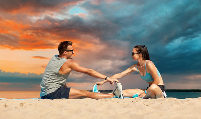 fitness, sport and lifestyle concept - smiling couple stretching legs on beach before training over sea and sunset sky on background
