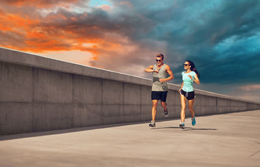 fitness, sport and lifestyle concept - happy couple in sports clothes and sunglasses running along concrete pier over sunset sky on background