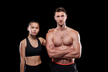 Fototapeta na wymiar Handsome muscular shirtless guy and his fit pretty girlfriend in activewear standing close to one another on dark background