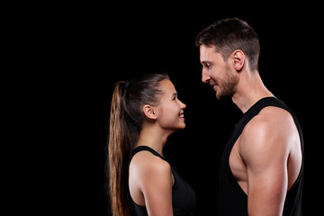 Fototapeta na wymiar Happy young sportswoman and sportsman in activewear looking at one another with smiles over black background
