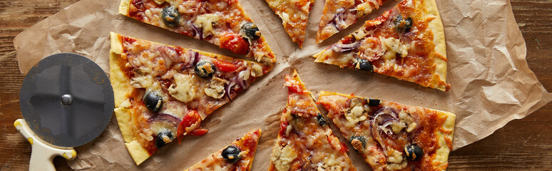 top view of cut delicious italian pizza with olives on baking paper near pizza knife on wooden background, panoramic shot