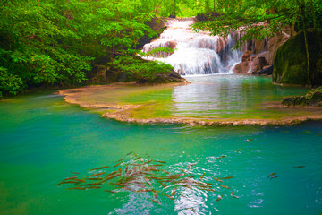 Fototapeta na wymiar Waterfalls and fish swim in the emerald blue water in Erawan National Park. Erawan Waterfall is a beautiful natural rock waterfall in Kanchanaburi, Thailand.Onsen atmosphere.