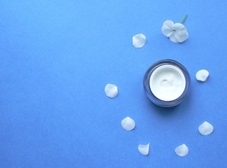 Glass jar of a beauty cream on a blue background, decorated with white flower petals. Concept of...