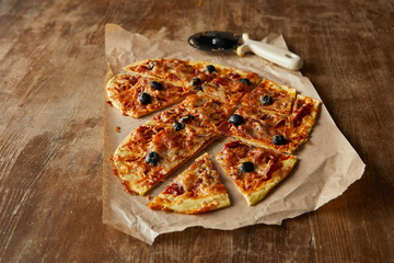 delicious italian pizza in heart shape cut into pieces on baking paper near pizza knife on wooden table