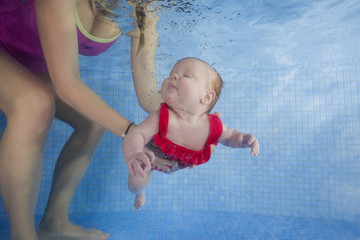 Newborn girl in a red swimsuit learns to dive in a swimming pool, mother holding the child.