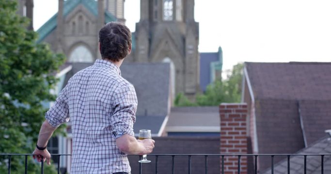 Trendy young man stands on balcony and looks off into cool skyline - traveling to nee city - wide shot