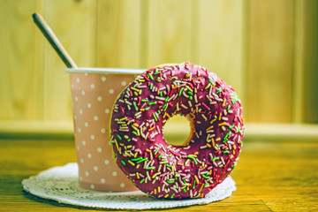 A paper cup of hot fresh brewed coffee next to a  pink icing donut on a wooden background, copy...