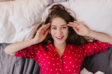 Fototapeta premium Amazing dark-haired female model enjoying morning in weekend. Indoor overhead photo of enthusiastic brunette girl lying in bed and laughing.