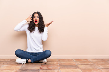 Young woman sitting on the floor making phone gesture and doubting