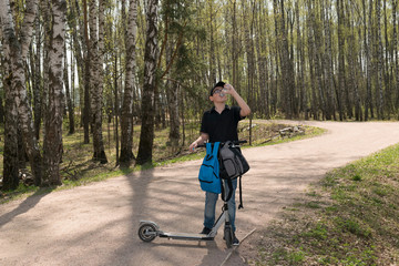 a teenager with a scooter stopped and drinks water from a bottle on a forest road, two backpacks...