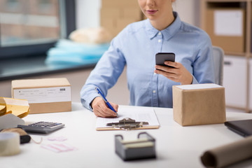 delivery, mail service, people and shipment concept - close up of woman with smartphone and parcel boxes filling checklist on clipboard at post office