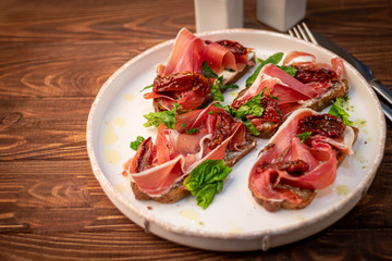 Sandwiches with prosciutto and sun-dried tomatoes on dark bread, spread with goat cheese. On a wooden background.