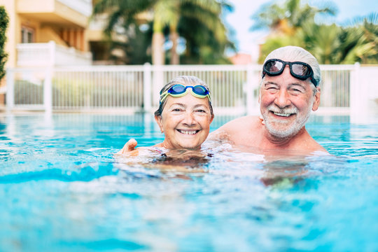 couple of cute seniors and pensioners in the water of the pool having fun and enjoying together - two mature people in love looking at the camera - doing exercise and training together smiling