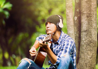 asian men wear a yarn hat, listen to headphones through Bluetooth, play guitar in the park, weekend