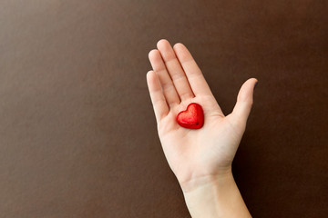 sweets, confectionery and food concept - hand holding heart shaped chocolate candy in red foil wrapper on brown background